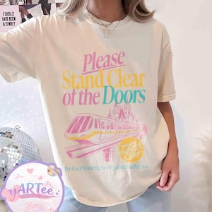 May include: Cream-colored t-shirt with the text "Please Stand Clear of the Doors" in pink, yellow, and teal. A pink and yellow monorail graphic is below the text, with a castle in the background. The text "Por Favor Mant&eacute;ngase Alejado de las Puertas" is below the graphic.