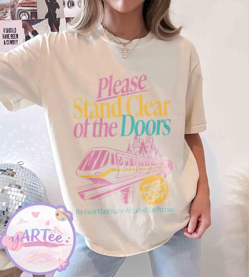 May include: Cream-colored t-shirt with the text "Please Stand Clear of the Doors" in pink, yellow, and teal. A pink and yellow monorail graphic is below the text, with a castle in the background. The text "Por Favor Mant&eacute;ngase Alejado de las Puertas" is below the graphic.