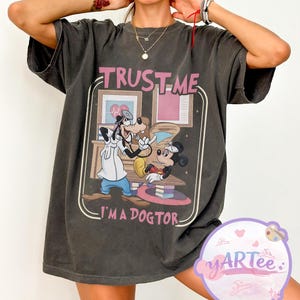 May include: Dark grey t-shirt with a graphic of Mickey Mouse, Goofy, and the text "TRUST ME I'M A DOGTOR." The design is framed with a light brown border. The shirt has short sleeves and a relaxed fit.