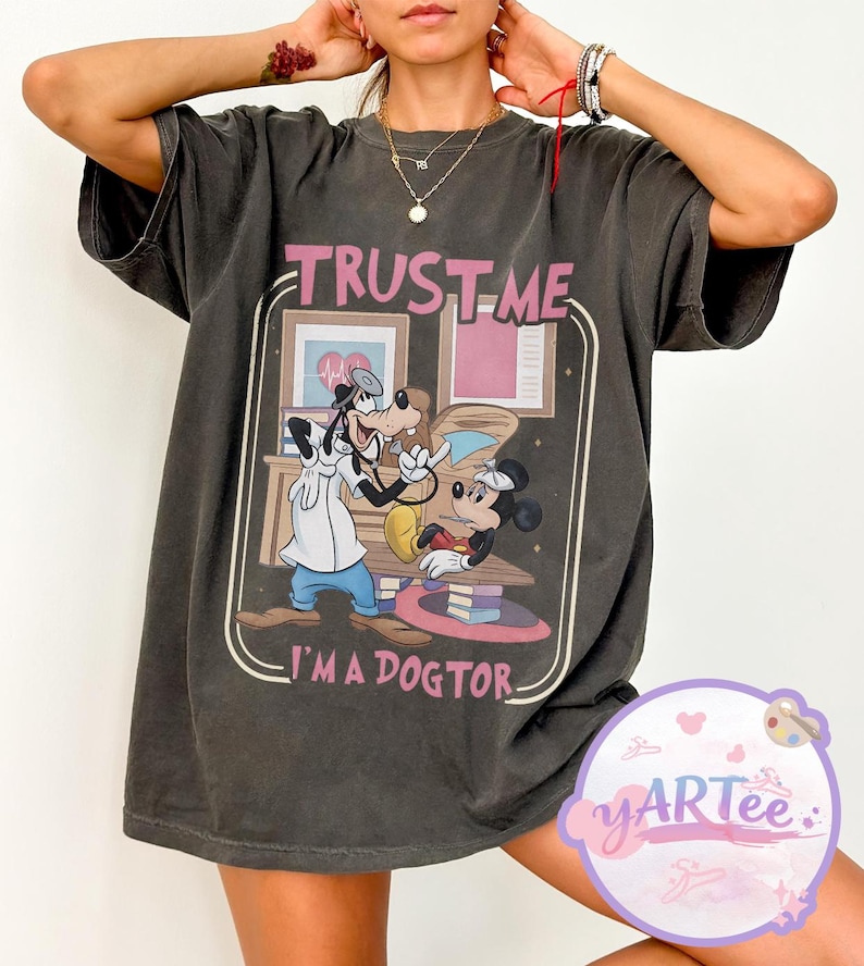 May include: Dark grey t-shirt with a graphic of Mickey Mouse, Goofy, and the text "TRUST ME I'M A DOGTOR." The design is framed with a light brown border. The shirt has short sleeves and a relaxed fit.