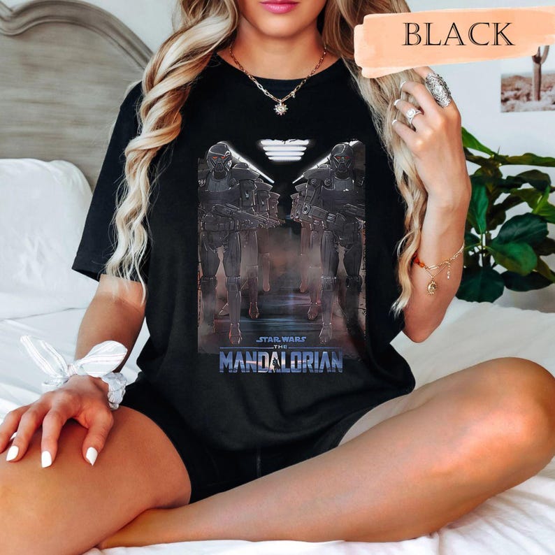 May include: Black t-shirt featuring a graphic from "Star Wars: The Mandalorian." The design includes two figures in dark armor, the show's logo, and the word "BLACK" in a peach-colored banner. The shirt is a casual wear item.