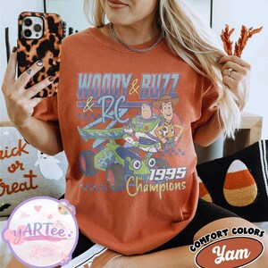 May include: Rust-colored t-shirt featuring a graphic of Buzz Lightyear and Woody from Toy Story, with the text "WOODY & BUZZ & RC 1995 Champions." The shirt is a Comfort Colors brand.