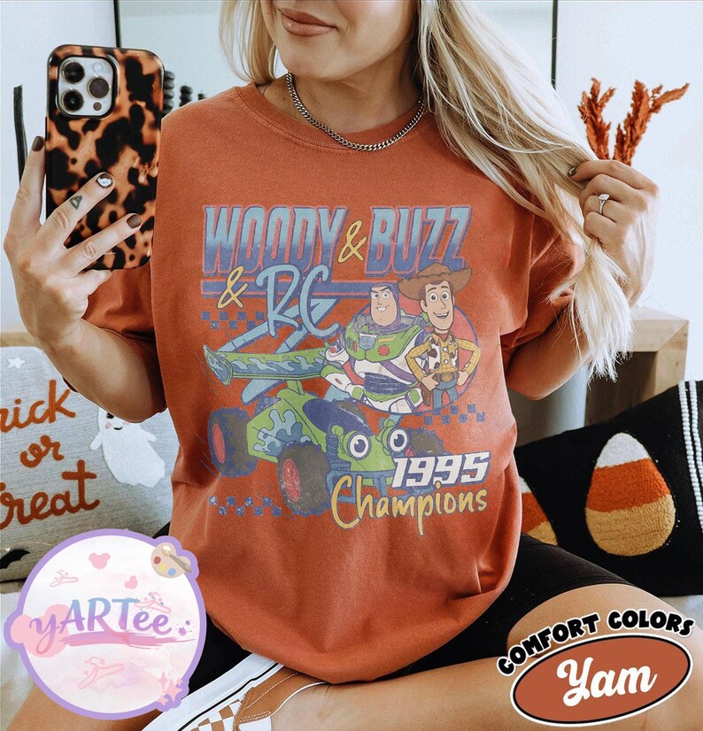 May include: Rust-colored t-shirt featuring a graphic of Buzz Lightyear and Woody from Toy Story, with the text "WOODY & BUZZ & RC 1995 Champions." The shirt is a Comfort Colors brand.