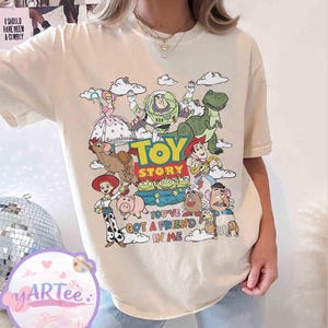 May include: Cream-colored t-shirt featuring a colorful graphic of characters from the animated film "Toy Story." The design includes the words "Toy Story" and "You've Got a Friend in Me."