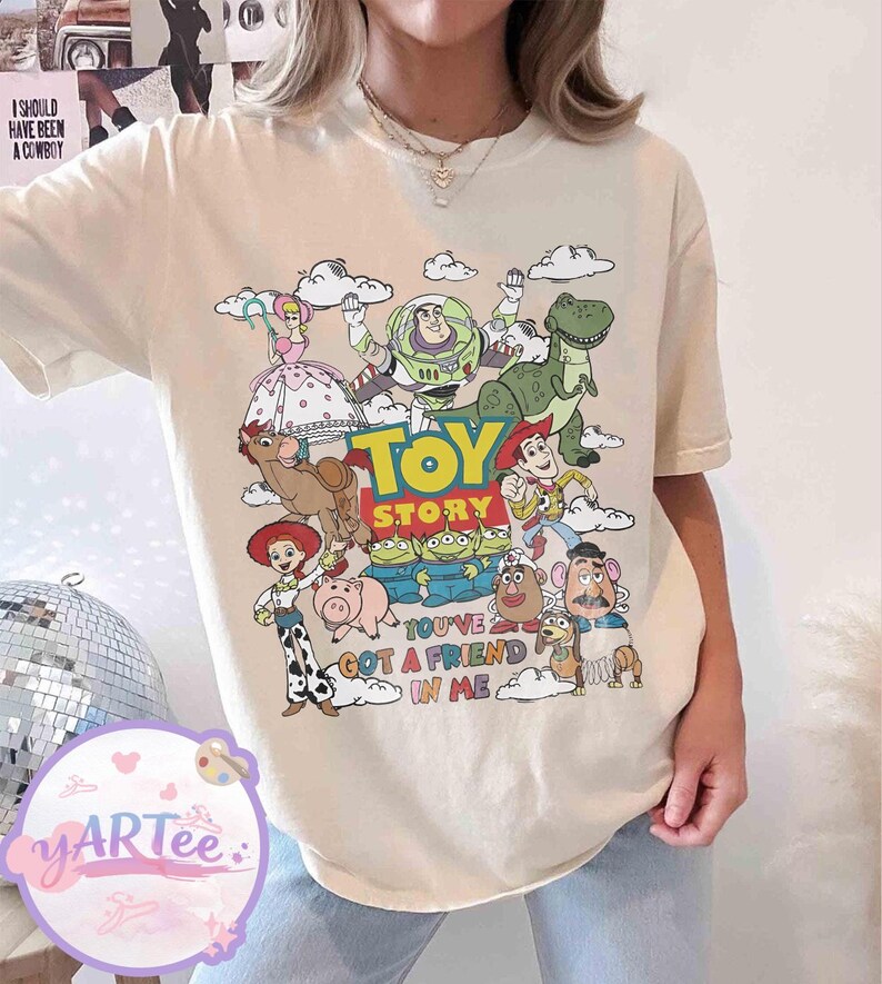 May include: Cream-colored t-shirt featuring a colorful graphic of characters from the animated film "Toy Story." The design includes the words "Toy Story" and "You've Got a Friend in Me."