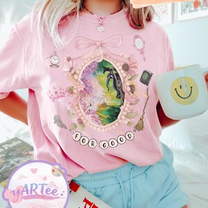 K&ouml;nnte beinhalten: Hellrosa T-Shirt mit einem gerahmten Fantasy-Motiv, das ein Pferd und die Worte "FOR GOOD" zeigt. Das Design beinhaltet eine Krone, einen Spiegel und eine Uhr. Eine wei&szlig;e Tasse mit einem gelben Smiley wird gehalten. Das Wort "BLOSSOM" ist ebenfalls sichtbar.