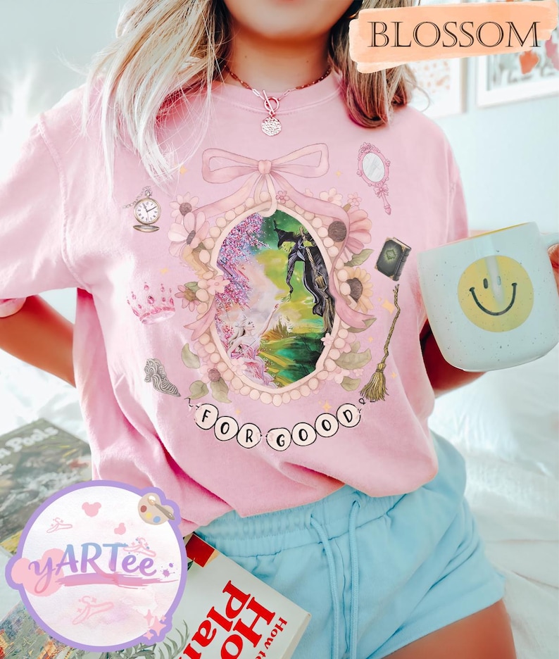 K&ouml;nnte beinhalten: Hellrosa T-Shirt mit einem gerahmten Fantasy-Motiv, das ein Pferd und die Worte "FOR GOOD" zeigt. Das Design beinhaltet eine Krone, einen Spiegel und eine Uhr. Eine wei&szlig;e Tasse mit einem gelben Smiley wird gehalten. Das Wort "BLOSSOM" ist ebenfalls sichtbar.