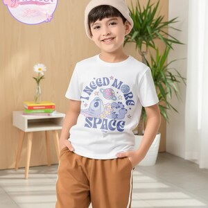 May include: A young person wearing a white t-shirt with the text "I need more space" and a graphic of a cartoon character, planets, and stars. The person is also wearing brown pants and a beige beanie.