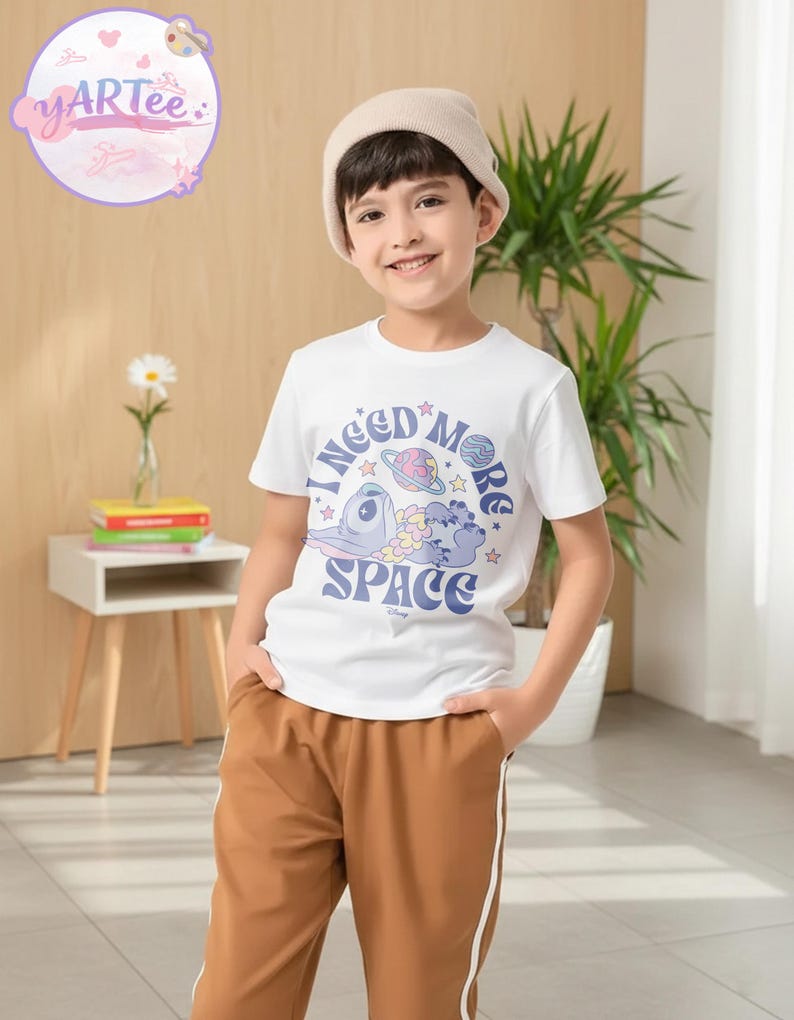 May include: A young person wearing a white t-shirt with the text "I need more space" and a graphic of a cartoon character, planets, and stars. The person is also wearing brown pants and a beige beanie.