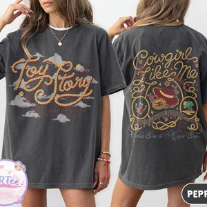 May include: A charcoal gray short-sleeve t-shirt featuring a front graphic with the text Toy Story in yellow rope lettering over gray clouds. The back displays the text Cowgirl Like Me and Takes One to Know One with a hat and rope design.