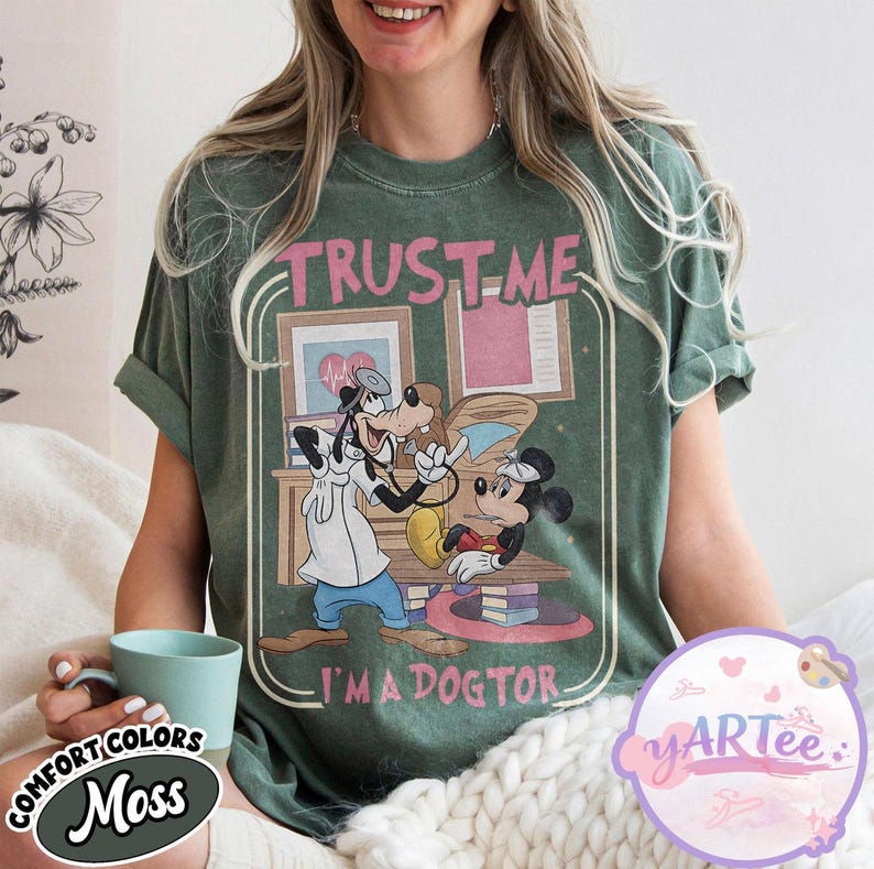 May include: Moss green t-shirt with a graphic of Goofy as a doctor examining Minnie Mouse. The text "TRUST ME I'M A DOGTOR" is printed above and below the cartoon characters. The shirt has rolled-up sleeves and a relaxed fit.