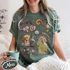 May include: Green t-shirt with a graphic featuring characters from "The Nightmare Before Christmas." The design includes Jack Skellington, Sally, Oogie Boogie, and other elements from the movie. The shirt has the text "COMFORT COLORS Moss".