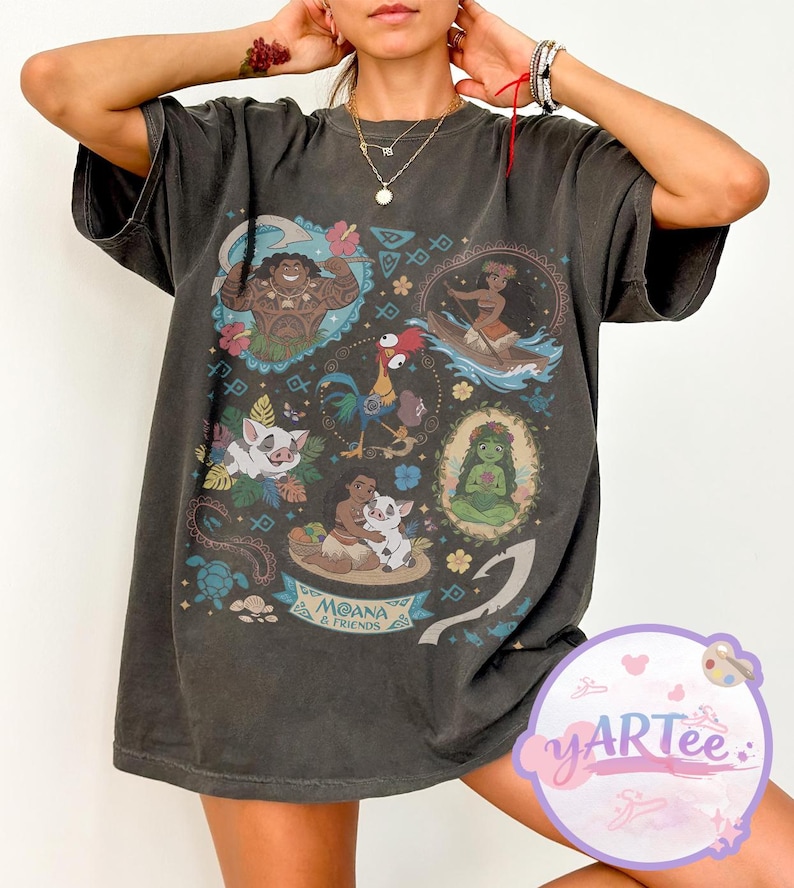 K&ouml;nnte beinhalten: Dunkelgraues T-Shirt mit einer farbenfrohen Grafik von Charakteren aus dem Animationsfilm "Vaiana". Das Design enth&auml;lt Bilder von Maui, Vaiana, Pua, Hei Hei und anderen Charakteren. Die Worte "Moana & Friends" sind ebenfalls sichtbar.