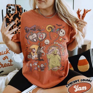 May include: Rust-colored t-shirt featuring a colorful graphic of characters and elements from "The Nightmare Before Christmas." The design includes Jack Skellington, Sally, and Oogie Boogie. The shirt has the words "Comfort Colors" and "Yam" printed on it.