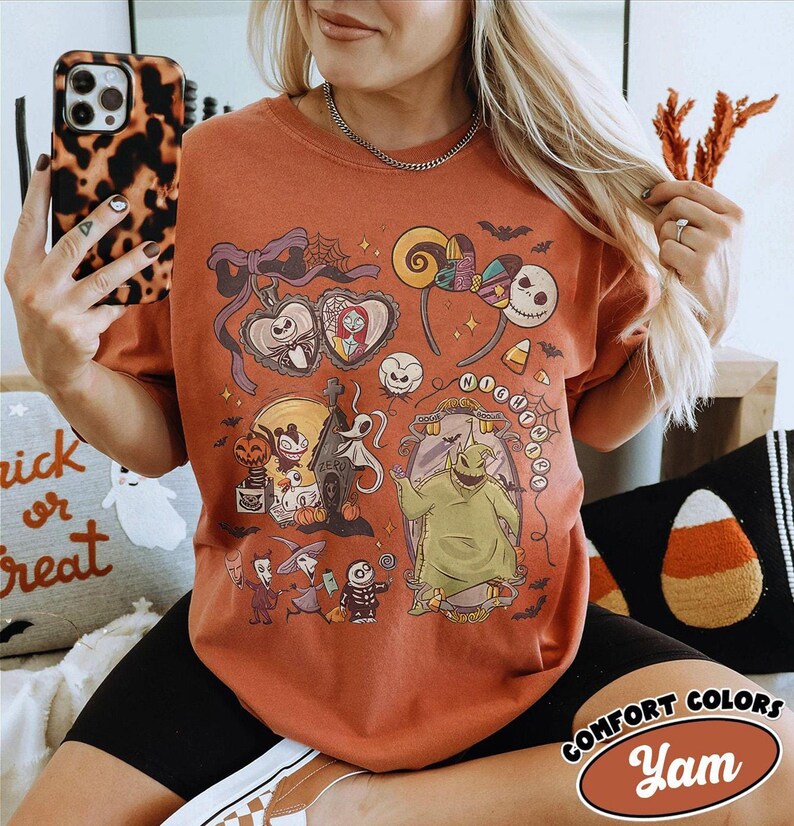 May include: Rust-colored t-shirt featuring a colorful graphic of characters and elements from "The Nightmare Before Christmas." The design includes Jack Skellington, Sally, and Oogie Boogie. The shirt has the words "Comfort Colors" and "Yam" printed on it.