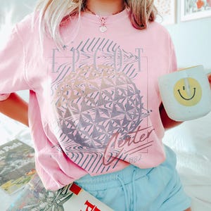 May include: A light pink t-shirt with a graphic of a geometric sphere and the text "EPCOT Center 1982". The shirt is paired with light blue shorts and a white mug with a yellow smiley face.