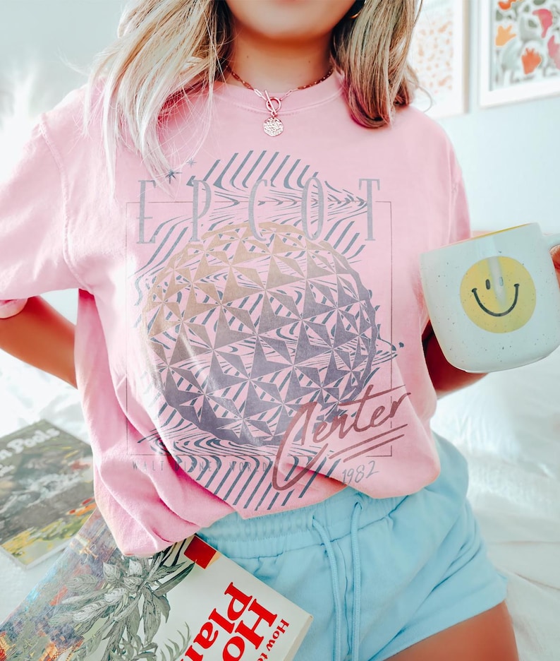May include: A light pink t-shirt with a graphic of a geometric sphere and the text "EPCOT Center 1982". The shirt is paired with light blue shorts and a white mug with a yellow smiley face.