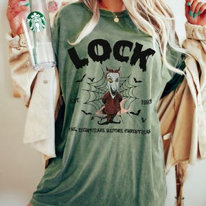 May include: Moss green t-shirt featuring a graphic of Lock from The Nightmare Before Christmas. The design includes the word "LOCK" above an illustration of the character, a spiderweb, bats, and the text "EST. 1993 THE NIGHTMARE BEFORE CHRISTMAS."