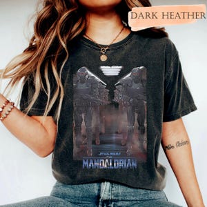 May include: Dark heather gray t-shirt featuring a graphic from Star Wars: The Mandalorian. The graphic shows two figures in black armor with red visors, holding weapons. The text "STAR WARS THE MANDALORIAN" is below the figures.