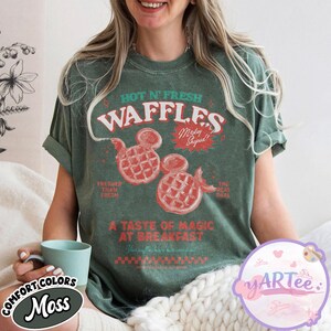 Puede incluir: Camiseta verde con un gr&aacute;fico que presenta el texto "HOT N' FRESH WAFFLES" y dos gofres con forma de Mickey Mouse. La camiseta tambi&eacute;n incluye las frases "Fresher Than Fresh" y "The Real Deal".