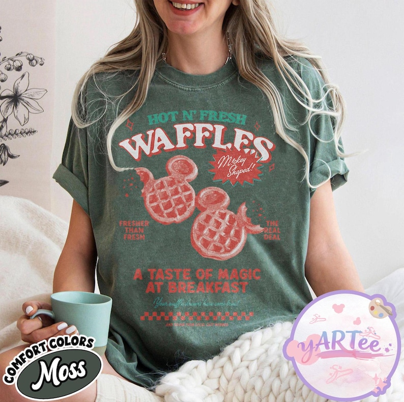 Puede incluir: Camiseta verde con un gr&aacute;fico que presenta el texto "HOT N' FRESH WAFFLES" y dos gofres con forma de Mickey Mouse. La camiseta tambi&eacute;n incluye las frases "Fresher Than Fresh" y "The Real Deal".