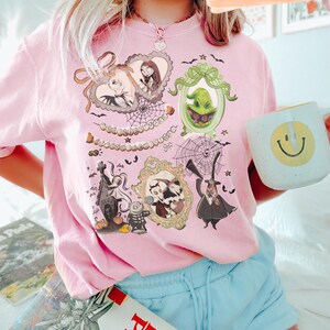 Puede incluir: Una camiseta rosa claro con ilustraciones enmarcadas de personajes de Pesadilla antes de Navidad. La camiseta tiene cuello redondo y mangas cortas. Una taza blanca con una carita sonriente amarilla se sostiene en la mano.