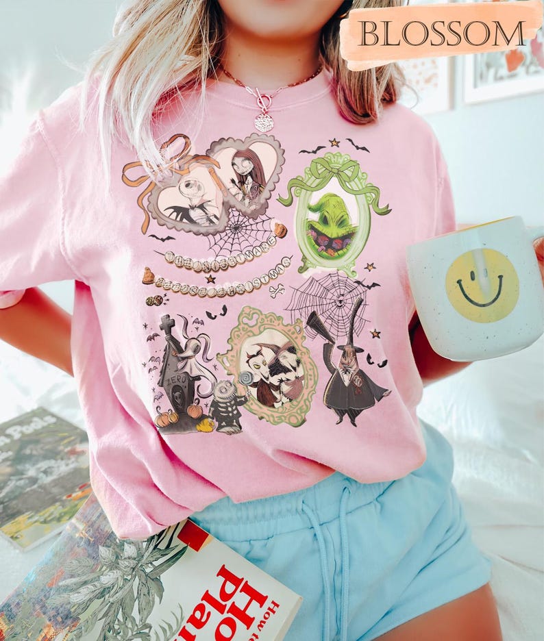 Puede incluir: Una camiseta rosa claro con ilustraciones enmarcadas de personajes de Pesadilla antes de Navidad. La camiseta tiene cuello redondo y mangas cortas. Una taza blanca con una carita sonriente amarilla se sostiene en la mano.