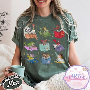 Puede incluir: Una camiseta verde musgo con ilustraciones de dibujos animados de personajes leyendo libros. Las ilustraciones incluyen personajes de cuentos populares. La camiseta tiene las palabras "COMFORT COLORS" y "Moss".