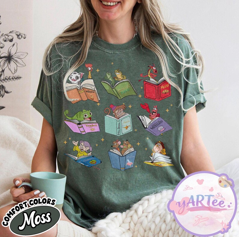 Puede incluir: Una camiseta verde musgo con ilustraciones de dibujos animados de personajes leyendo libros. Las ilustraciones incluyen personajes de cuentos populares. La camiseta tiene las palabras "COMFORT COLORS" y "Moss".