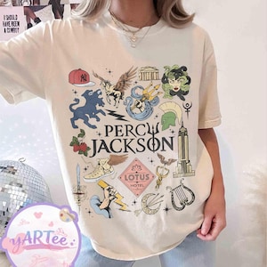 Pu&ograve; includere: T-shirt color crema con un design grafico colorato e la scritta "Percy Jackson". Il design include illustrazioni di creature mitologiche, simboli e il logo Lotus Hotel. Stile casual e unisex.