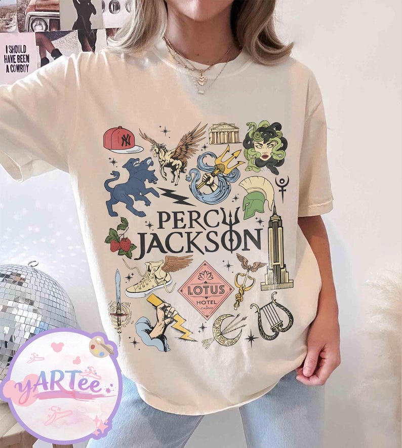Pu&ograve; includere: T-shirt color crema con un design grafico colorato e la scritta "Percy Jackson". Il design include illustrazioni di creature mitologiche, simboli e il logo Lotus Hotel. Stile casual e unisex.