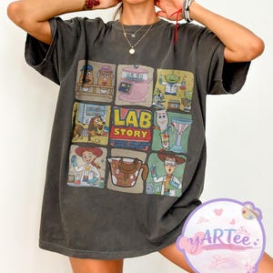 May include: Dark grey t-shirt featuring a colorful graphic of cartoon characters in a lab setting. The graphic includes the text "LAB STORY" and other text. The shirt is a casual, oversized style.