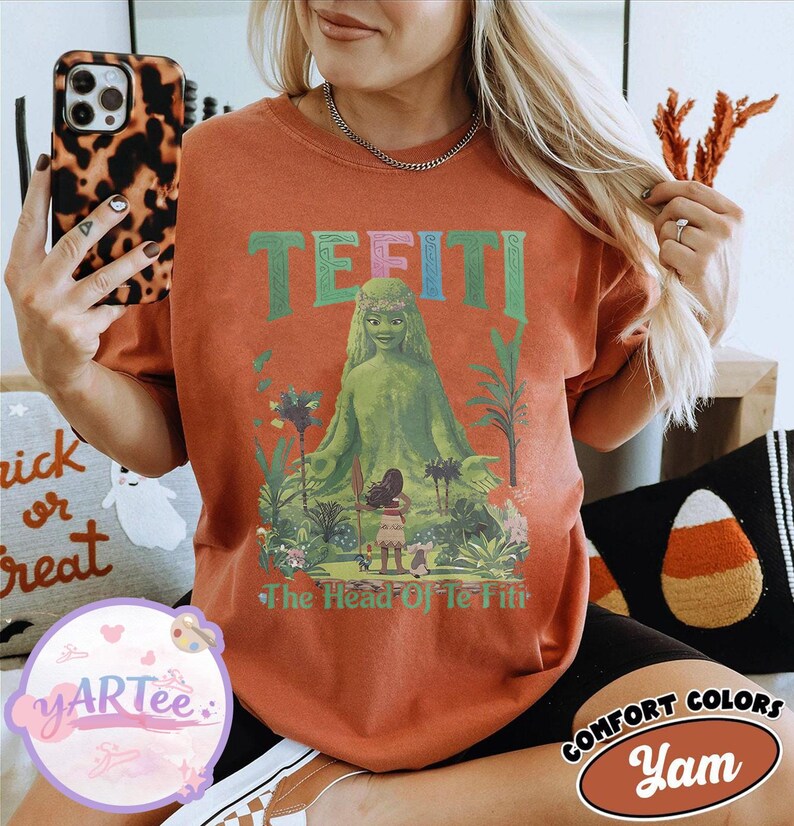 K&ouml;nnte beinhalten: Ein orangefarbenes T-Shirt mit einem Motiv von Te Fiti aus dem Film Moana. Das Design enth&auml;lt das Wort "TE FITI" in Gr&uuml;n und Blau sowie den Satz "The Head Of Te Fiti". Das Shirt geh&ouml;rt zur Comfort Colors Kollektion.