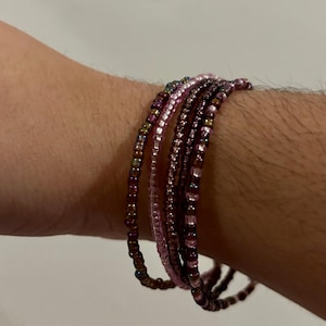 Puede incluir: Un conjunto de cinco pulseras de cuentas en tonos rosa y marrón. Las pulseras están apiladas en la muñeca de una persona.