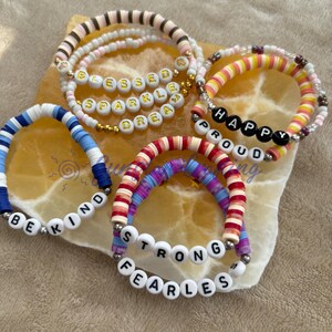 Puede incluir: Un conjunto de pulseras de cuentas coloridas con mensajes positivos, que incluyen "Blessed", "Sparkle", "Dream", "Happy", "Proud", "Be Kind", "Strong" y "Fearless".