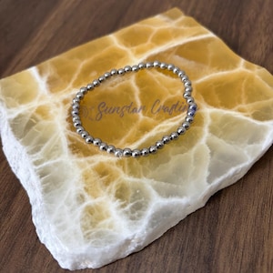 May include: A silver beaded bracelet with a single clear crystal bead. The bracelet is on a yellow and white stone surface.
