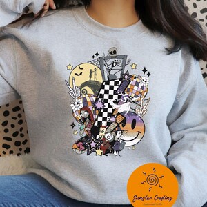 May include: A light gray sweatshirt featuring a colorful Halloween-themed graphic. The design includes a checkered pattern, a smiling face, stars, and characters from The Nightmare Before Christmas. The sweatshirt has a crew neck.