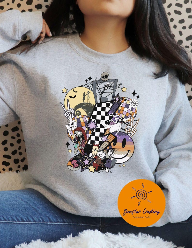 May include: A light gray sweatshirt featuring a colorful Halloween-themed graphic. The design includes a checkered pattern, a smiling face, stars, and characters from The Nightmare Before Christmas. The sweatshirt has a crew neck.