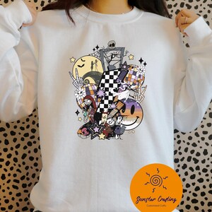 May include: White sweatshirt featuring a colorful graphic design with Halloween and Nightmare Before Christmas themes. The design includes a checkered pattern, a smiley face, stars, and various characters. The sweatshirt has long sleeves and a crew neck.
