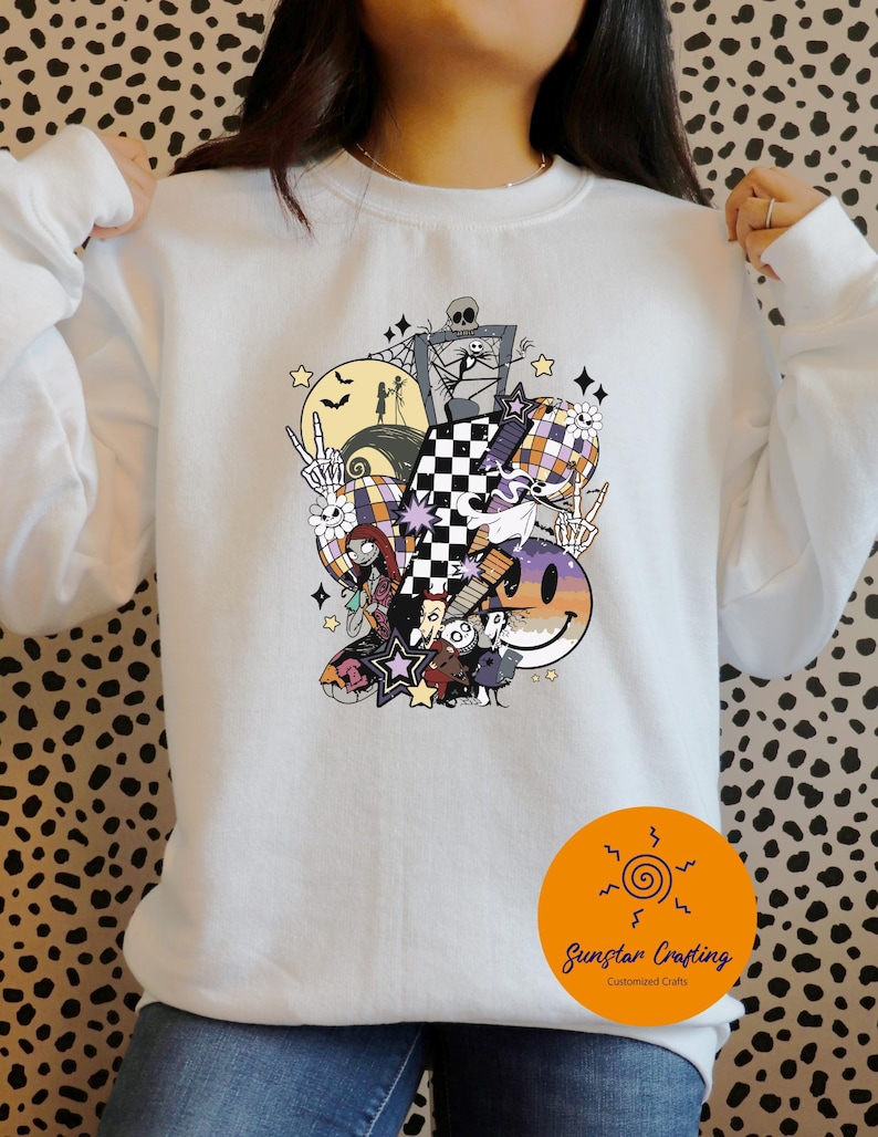 May include: White sweatshirt featuring a colorful graphic design with Halloween and Nightmare Before Christmas themes. The design includes a checkered pattern, a smiley face, stars, and various characters. The sweatshirt has long sleeves and a crew neck.