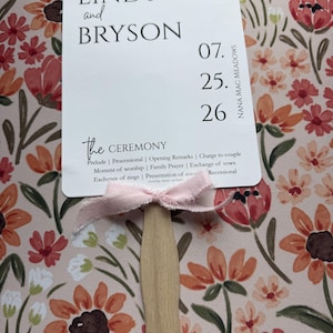 May include: A white rectangular wedding program fan with rounded corners, featuring the text Welcome to our wedding, Lindy and Bryson, 07.25.26, Nana Mac Meadows, and The Ceremony. A light pink frayed ribbon is tied to the wooden handle.