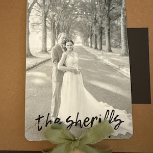 May include: Black and white photograph of a couple on a tree-lined road, likely a wedding photo. The photo is mounted on a brown background with a green ribbon and the text "the sherilly".
