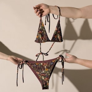Może przedstawiać: Dwuczęściowe bikini z wzorem grzybów i kwiatów. Trójkątny top i klasyczne figi są w kolorze bordowym z wzorem kolorowych grzybów i kwiatów. Bikini ma ramiączka i wiązane boki.