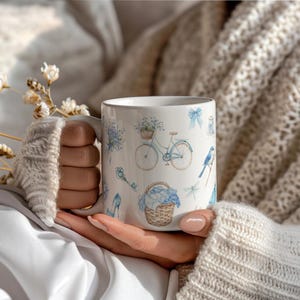 May include: White ceramic mug with a blue and white floral design. The mug features a bicycle with a basket of flowers, a key, a blue bird, a dragonfly, and a blue shoe.