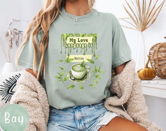 以下が含まれることがあります： 「My Love Language is Matcha」の文字と抹茶ラテのイラストが描かれた、淡い緑色のTシャツ。デザインには緑の葉とソーサーに乗った抹茶のカップが含まれています。ゆったりとしたフィット感のシャツです。