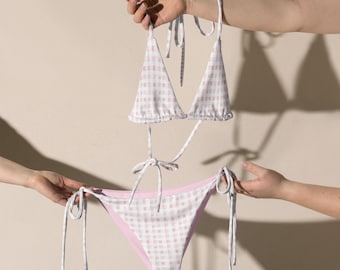 May include: A two-piece bikini set with a pink and white gingham pattern. The top is a triangle style with tie straps, and the bottoms have tie sides. The bikini is held up by hands against a neutral background.