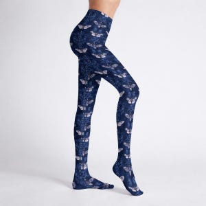 May include: Navy blue leggings with a repeating pattern of white moths and ornate floral designs. The leggings are high-waisted and form-fitting, made of a stretchy material. The overall aesthetic is dark and gothic.