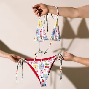 May include: A two-piece bikini set with a white base and a colorful pattern of cherries, strawberries, and floral designs. The bikini top has triangle cups and adjustable ties. The bottoms are red-lined with matching ties.