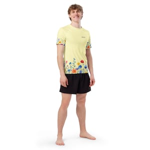 Pode incluir: Uma camisa amarela clara de manga curta com um design floral na parte inferior e nas mangas. A camisa tem um pequeno logotipo na parte superior do peito e é combinada com calções pretos. O design floral inclui flores vermelhas, azuis e amarelas.