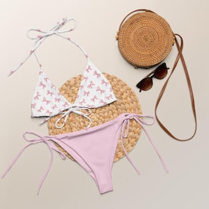 May include: A white bikini top with pink bow accents and a matching light pink bikini bottom. A round woven bag, sunglasses, and a woven placemat are also in the image. The bikini is a summer swimwear item.